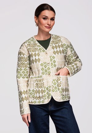 Kobieta nosząca zieloną i beżową wzorzystą zapinaną na guziki kurtkę z przednimi kieszeniami na ciemnozielonym topie i ciemnoniebieskich dżinsach.