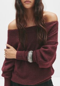 Bordeaux off-shoulder trui met een textuur gebreid, voorzien van geribbelde manchetten. Een brede zwarte armband met zilveren studs siert de pols.
