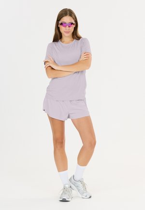 Jonge vrouw draagt lichtpaarse sportshorts en T-shirt, witte sokken, zilveren sneakers en paars getinte sportzonnebril, staand met gekruiste armen.