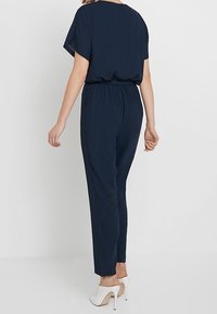 Mörkblå jumpsuit med korta ärmar, elastisk midja och smalnande ben; har ett glatt tyg och en avslappnad passform, matchad med vita klackar.