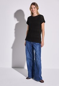 Svart stickad t-shirt med korta ärmar och texturerat mönster, kombinerad med vida blå denimjeans. Mörka slip-on skor fulländar looken.