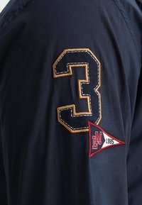 Camiseta azul marino con un gran número "3" dorado bordado y un parche triangular rojo con "LM 1985" en detalles negros y blancos.