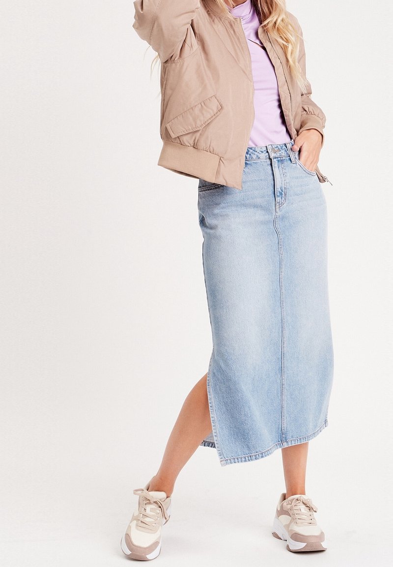Femme portant une veste beige, un haut lilas, une jupe midi en denim bleu clair avec une fente latérale, et des baskets blanc-beige, debout devant un fond uni.