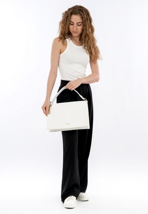 Woman with long curly hair wearing white tank top, black wide-leg pants, white sneakers, holding white handbag with gold accents.