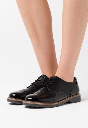 Czarne skórzane buty z lakierowanym wykończeniem, z wyróżniającymi się wzorami brogu, sznurowanym stylem oraz płaską podeszwą z brązowym akcentem na krawędzi.