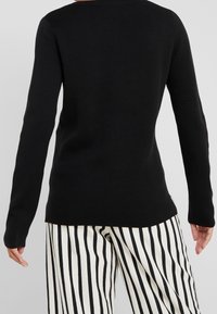 Pull en tricot noir à manches longues avec un col rond. Porté avec un pantalon rayé vertical noir et blanc. Texture lisse, style ajusté.