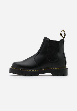 Botas Chelsea de cuero negro con paneles laterales elásticos, acentos de costura amarilla y suelas de goma texturizada. Presenta una pestaña en el talón.