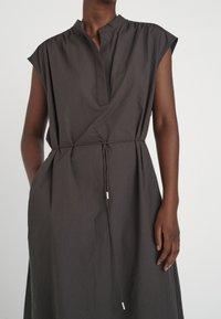 Ärmlös mörkgrå klänning med en smal V-ringning och tunn knytbelte vid midjan, bärs av en person med mörk hud mot en enfärgad bakgrund.