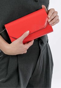 Clutch de imitación cuero rojo con acabado brillante, con tapa estilo sobre y correa de cadena en tono plateado, sostenido en una mano contra una tela oscura.