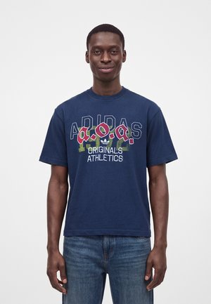 Junger Mann, der ein dunkelblaues Adidas Originals Athletics T-Shirt und blaue Jeans trägt, vor einem einfachen weißen Hintergrund stehend.