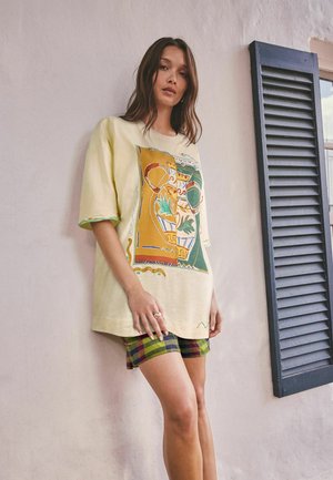 Femeie purtând un tricou bej supradimensionat cu design abstract colorat și pantaloni scurți în carouri, stând lângă un perete pal cu obloane întunecate la fereastră.