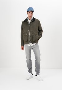 Green canvas jacket with a black collar, two front pockets, worn over a white shirt; paired with gray jeans and white sneakers. Blue cap atop.