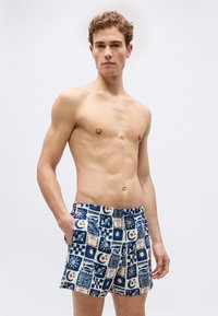 Άνδρας χωρίς πουκάμισο με κατσαρά μαλλιά, φορώντας μοτίβο μπλε και λευκά μαγιό με ναυτικά και ουράνια σύμβολα, στέκεται μπροστά σε μια απλή φόντο.