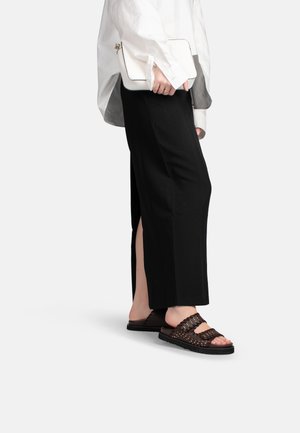 Persoon draagt zwarte wijde broek met zijsplit, witte oversized blouse, houdt een witte clutch vast en bruine open sandalen met studs.