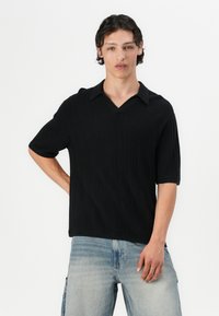 Črna rebrasta pletena majica s kratkimi rokavi in V-izrezom ter zloženo ovratnico. Oblečena je z svetlo modrimi poškodovanimi denim hlačami. Model pozira z eno roko na boku.