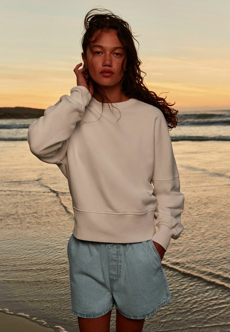 Ung kvinde med krøllet hår iført en hvid sweatshirt og denimshorts står på en strand ved solnedgang med bølger i baggrunden.