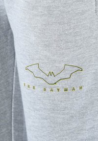 Graue Jogginghose mit einem Fledermaus-Logo und dem Text "THE BATMAN" in Gelb. Der Stoff hat eine weiche, strukturierte Optik.