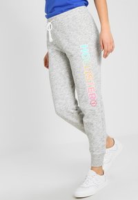 Ljusgrå joggers har en avslappnad passform, elasticitet i midjan med snörning och flerfärgad text "HOLLISTER" på vänster ben.
