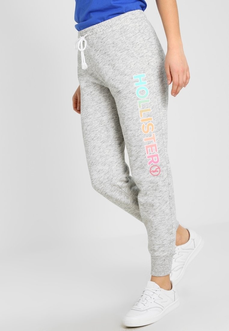 Ljusgrå joggers har en avslappnad passform, elasticitet i midjan med snörning och flerfärgad text "HOLLISTER" på vänster ben.