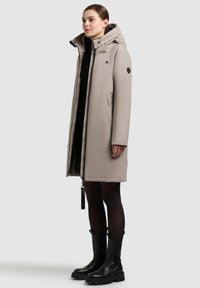 Béžová vypchatá parka s kapucňou, bočnými vreckami a predným zipsom. Doprevádza ju čierna robustná obuv. Hladká textúra, minimalistický dizajn.