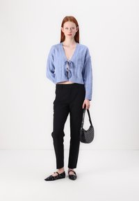 Hele helesinine lühike nööridega kaeluse-sweater, millel on V-kaelus, koos mustade kitsaste pükste ja mustade nurgeliselt kaunistatud sussidega, hoides musta kotti.