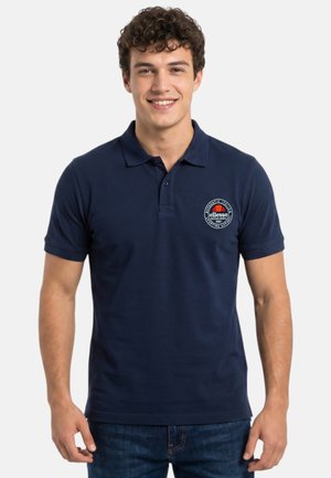Jeune homme aux cheveux bouclés portant un polo bleu marine avec un logo rond sur la poitrine gauche et un jean bleu, souriant.