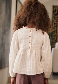 Children's white textured blouse with long sleeves, gathered waist, and five brown buttons on the back. Styled with pink pants.