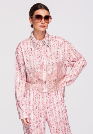 Nainen pukeutuneena vaaleanpunaiseen ja valkoiseen kuvioituun napilliseen paitaan ja housuihin, koristellut kaulus, pitsinen korsettivyö, suuret aurinkolasit ja renkaan malliset korvakorut.