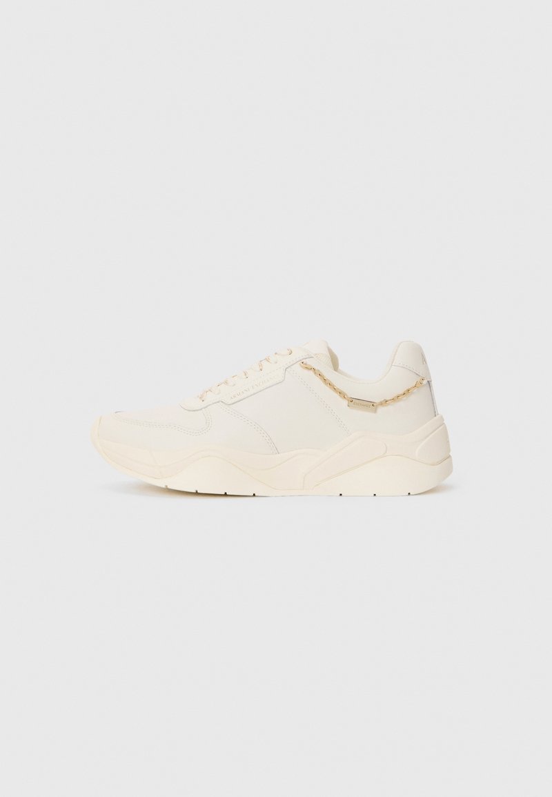 Cream-colored sneaker with a smooth leather upper, textured accents, stitched details, and a chunky rubber sole. Features a small decorative chain.