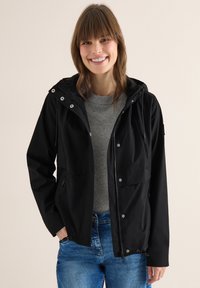 Femme souriante portant une veste à capuche noire, un pull gris et un jean bleu, debout devant un fond clair uni.