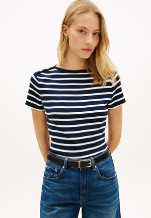 Šviesiaplaukė moteris, dėvinti auksinius lankelius ausyse, tamsiai mėlyną ir baltą dryžuotą trumpomis rankovėmis palaidinę su auksiniais mygtukais, mėlynas džinsus ir juodą diržą su auksine sagtimi.