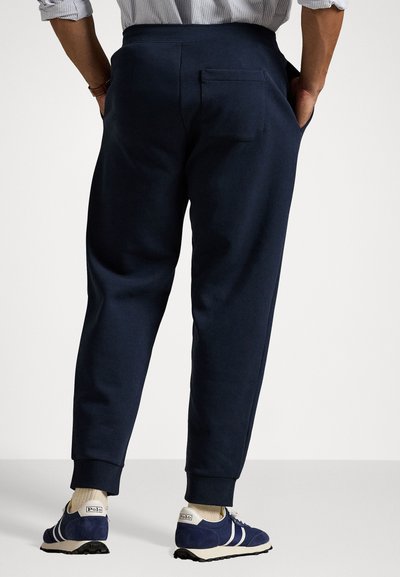 Navy blue joggers with elastic waistband and cuffed ankles. Features a back pocket. Worn with blue athletic shoes and light-colored socks.
