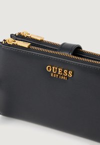 Bolsa sintética negra com fechos dourados, apresentando o logótipo em relevo "GUESS EST 1981". Textura suave e forma retangular com compartimentos duplos.