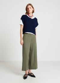 Marineblauer Strickpullover mit Zopfmuster, darüber ein weißes T-Shirt, kombiniert mit olivgrünen, weit geschnittenen Hosen und schwarzen Ballerinas mit dekorativen Metallakzenten.