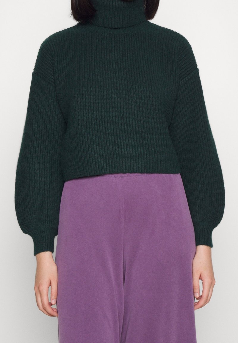 Dunkelgrüner, gerippterRollkragenspullover mit langen, ballonförmigen Ärmeln. Kombiniert mit hochgeschnittenen, helllila Hosen. Weiche Textur, cropped Design.