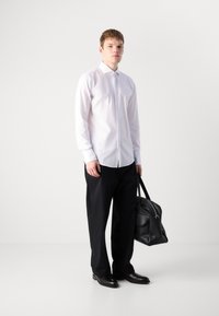 Witte lange mouw shirt gecombineerd met zwarte, losse broek en zwarte leren schoenen. De persoon houdt een zwarte leren tas vast.