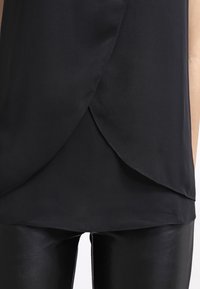 Blusa nera con un design a strati e avvolgente sul davanti. Il tessuto è liscio e leggermente trasparente, abbinato a leggings neri aderenti.