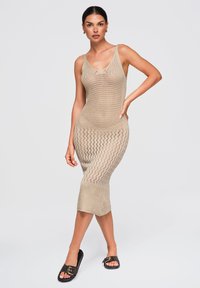 Femme debout, la main sur la hanche, portant une robe midi beige en maille sans manches et des sandales noires à boucle sur un fond gris clair.