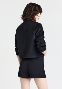 Giacca nera corta in tessuto testurizzato, con polsini a coste e schiena liscia. Abbinata a pantaloni corti neri aderenti, con un design minimalista.
