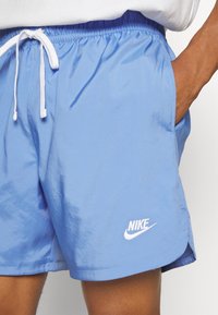 Detailní záběr na osobu v světle modrých Nike šortkách s bílou šňůrkou, ruka v kapse a viditelné logo Nike poblíž lemování.