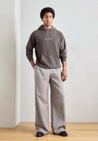 Bruine hoodie met voorzak en witte tekst, gecombineerd met lichtgrijze wijde broek en zwarte schoenen, staand op een houten vloer.