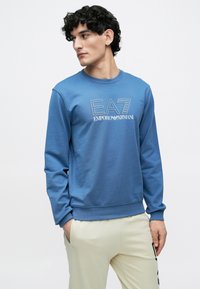 Zils džemperis ar "EA7 Emporio Armani" logotipu, apaļš kakls, garas piedurknes, ribota aproces un brīvs piegriezums, kombinēts ar bēšām biksēm.