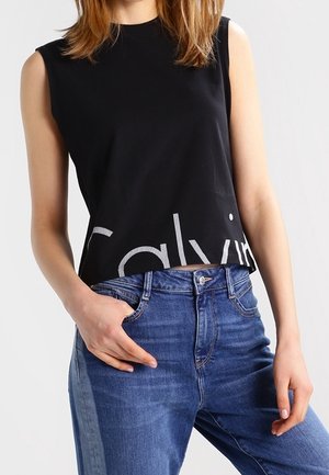 Kobieta nosząca czarny krótki top bez rękawów z częściowo widocznym białym napisem oraz wysokie niebieskie dżinsy z jedną ręką w kieszeni.