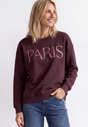 Kvinde med blondt hår smilende, iført en burgunder sweatshirt med ordet "PARIS" og blå jeans, hånden i lommen mod hvid baggrund.
