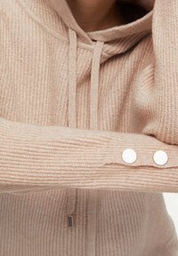 Hoodie beige en maille avec texture côtelée, cordons de serrage et deux boutons ronds blancs au niveau du poignet.