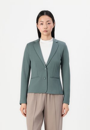 Fitted green blazer with a sleek design, two front pockets, and a single button closure, worn over a white high-neck top.