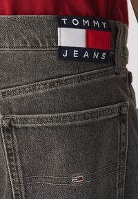 Pantalones de mezclilla gris con un bolsillo trasero que presenta un parche de logo de Tommy Jeans en azul marino, blanco y rojo, y un pequeño detalle de bandera bordada.
