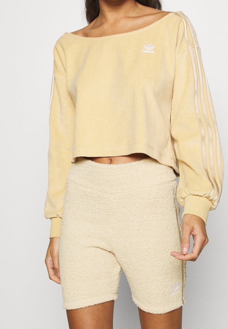 Sweatshirt jaune court avec design dénudé d'épaule, présentant trois rayures verticales sur les manches. Short beige texturé avec taille élastique.