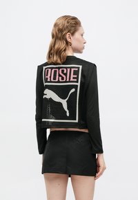 Schwarze, ärmellose Netz-Weste mit "ROSIE" in Pink und einer weißen Silhouette eines Pumas auf dem Rücken. Getragen über einem figurbetonten Langarmoberteil und einem Rock.