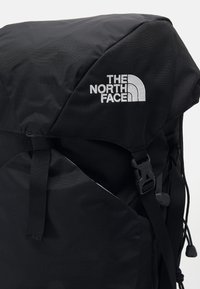 Schwarzer Stoffrucksack mit dreieckiger Oberseite, verstellbaren Trägern, Seitentaschen und einem gestickten Logo auf der Vorderseite. Strukturierter Material.